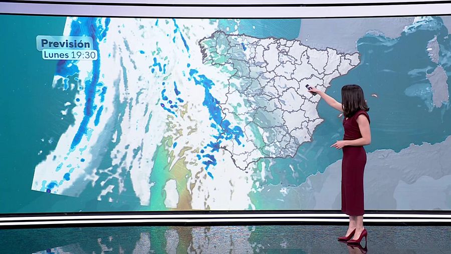 Una mujer, vestida de rojo, presenta el pronóstico del tiempo frente a una pantalla con un mapa de España y la previsión meteorológica, señalando un punto específico. En la esquina superior izquierda se lee 