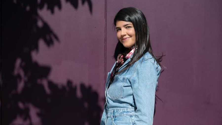 Una dona amb cabells foscos i llisos, vestida amb roba vaquera i un mocador al coll, posa davant d'una paret morada amb l'ombra d'un arbre projectada.