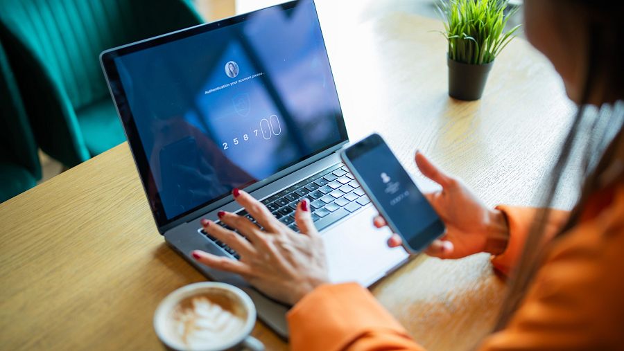Una mujer sentada en una mesa de una cafetería con un móvil en la mano derecha y la izqujierda sobre un el teclado de su ordenador portátil. En la pantalla se ven números similares a un código de verificiación. A su lado, una taza con un café