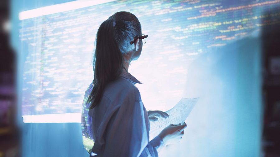 Una mujer de espaldas mirando una gran pantalla donde hay código de programación. En sus manos tiene un folio