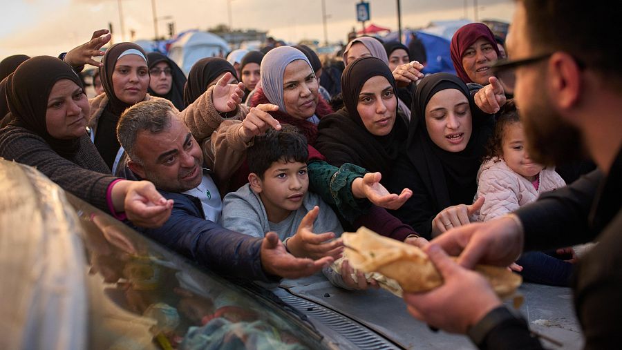 En un campo al aire libre, un hombre entrega pan a un grupo de personas, entre ellas mujeres con pañuelos y niños, que extienden sus manos en busca de alimento. La escena sugiere una situación de necesidad.