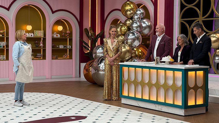 En un plató decorado en tonos rosas y dorados, Belén Esteban, con camisa azul y delantal blanco, se enfrenta a los jueces de un programa culinario, incluyendo una jueza con un traje dorado.