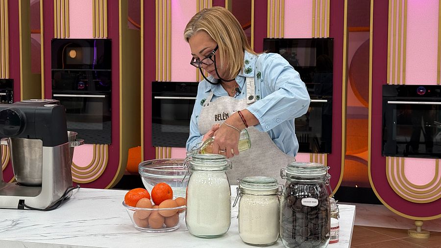 En un plató de televisión, Belén Esteban, con gafas y delantal, cocina. Se observan ingredientes como huevos y naranjas, junto a tarros y una batidora, con un fondo decorado con paneles y hornos.