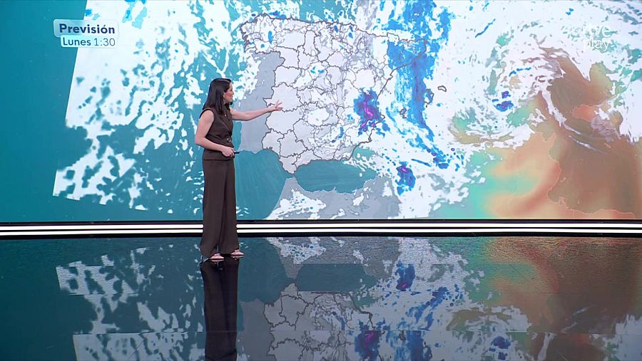 Una presentadora de televisión, vestida con un conjunto marrón, presenta el pronóstico del tiempo frente a una pantalla que muestra un mapa de España con información meteorológica. La presentadora señala una zona del mapa, mientras que en la pantalla se lee la previsión para el lunes.