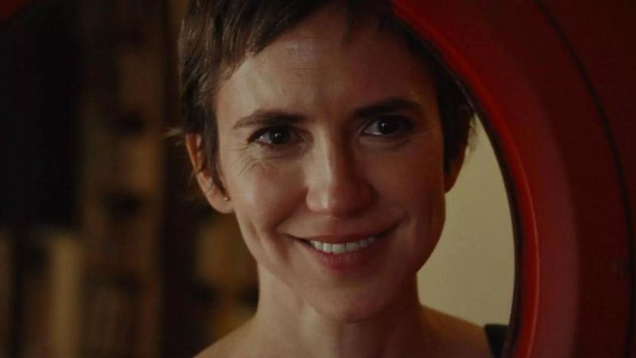 En primer plano, Aina Clotet sonríe con el pelo corto castaño, enmarcada parcialmente por un elemento circular rojo, con un fondo borroso que sugiere un interior.