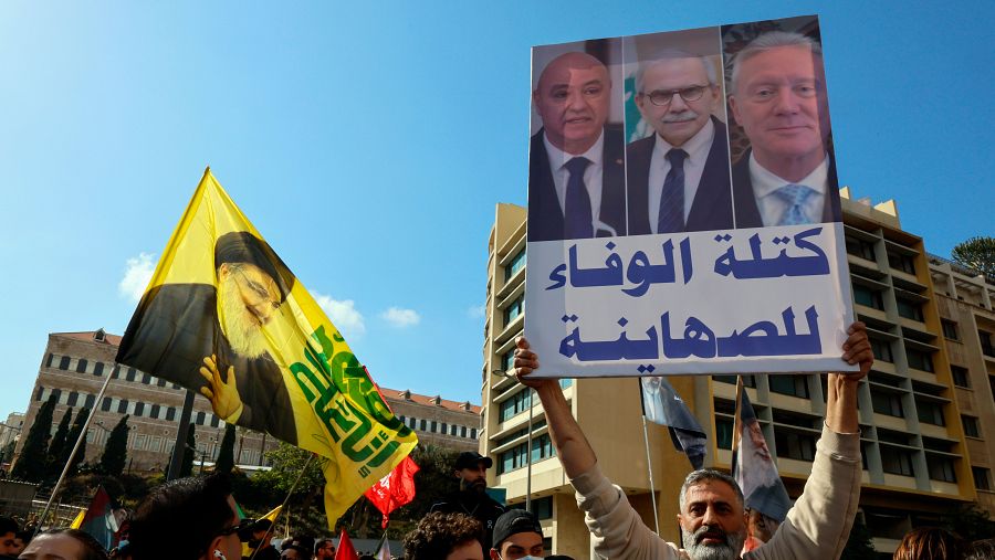 En Beirut, durante una manifestación, un hombre sostiene un cartel con fotos y texto en árabe. Una bandera amarilla con una figura religiosa se alza sobre la multitud, mientras que los manifestantes se agrupan frente a edificios.