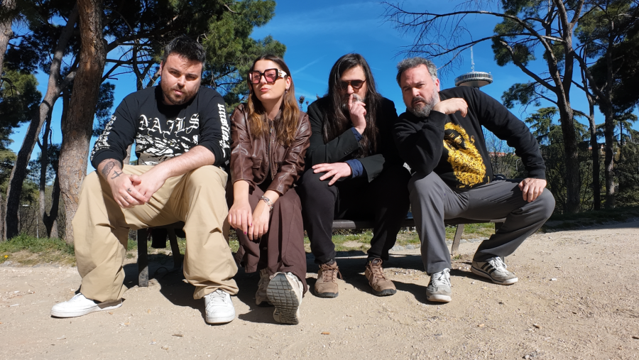 Cuatro personas se encuentran sentadas en un banco al aire libre, con árboles y una torre de telecomunicaciones como telón de fondo. Los individuos visten ropa casual, como camisetas, chaquetas y sudaderas.
