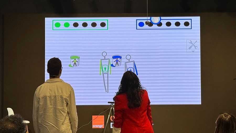En primer término hay dos personas, un joven y una mujer de espaldas, que miran a una proyección done ven dos pictogramas y un par de dibujos de cubos llenos de pintura