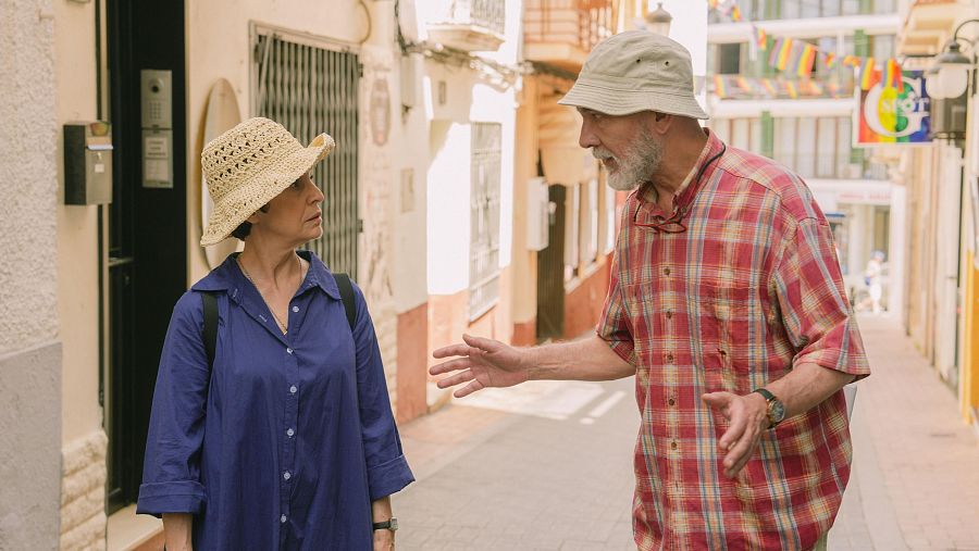 En una calle mediterránea, un hombre con sombrero tipo safari y camisa de cuadros conversa con una mujer que lleva un sombrero de paja y camisa azul, ambos turistas en un entorno urbano con edificios blancos y detalles coloridos.