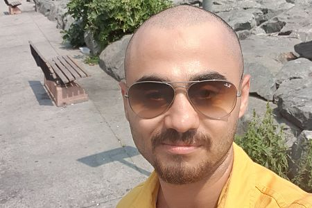 Hombre con cabeza rapada, bigote y barba corta, viste camisa amarilla y gafas Ray-Ban. Fondo con camino, banco y rocas.