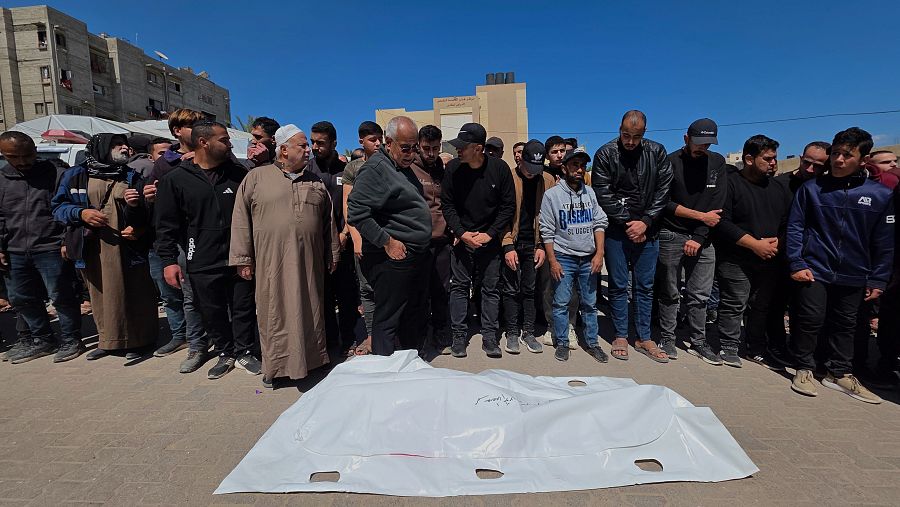 Hombres de diversas edades se reúnen en Gaza alrededor de un cuerpo cubierto con un sudario blanco, en un acto de despedida. La escena muestra una mezcla de vestimentas tradicionales y modernas.