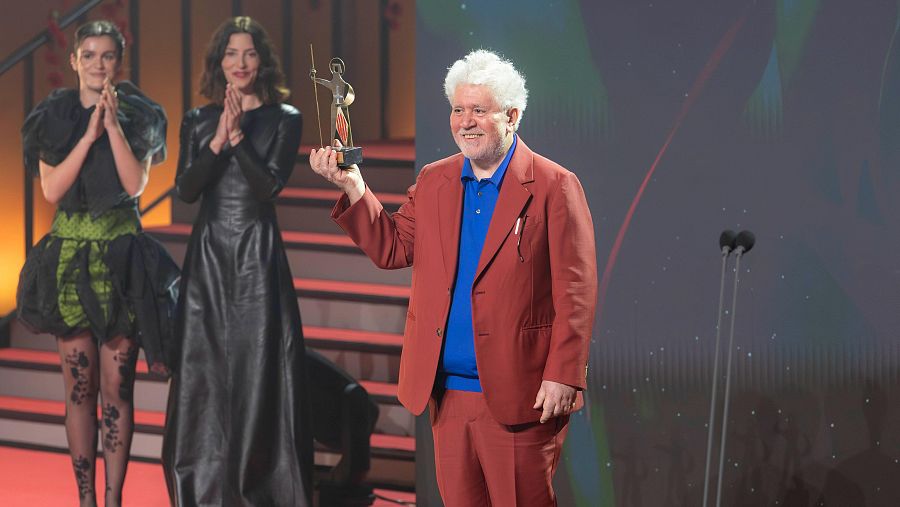 Pedro Almodóvar recibe un premio dorado de manos de Bárbara Lennie y Amaia, quienes aplauden. Almodóvar viste traje teja y polo azul; Lennie, vestido negro; Amaia, vestido negro y verde con medias florales.