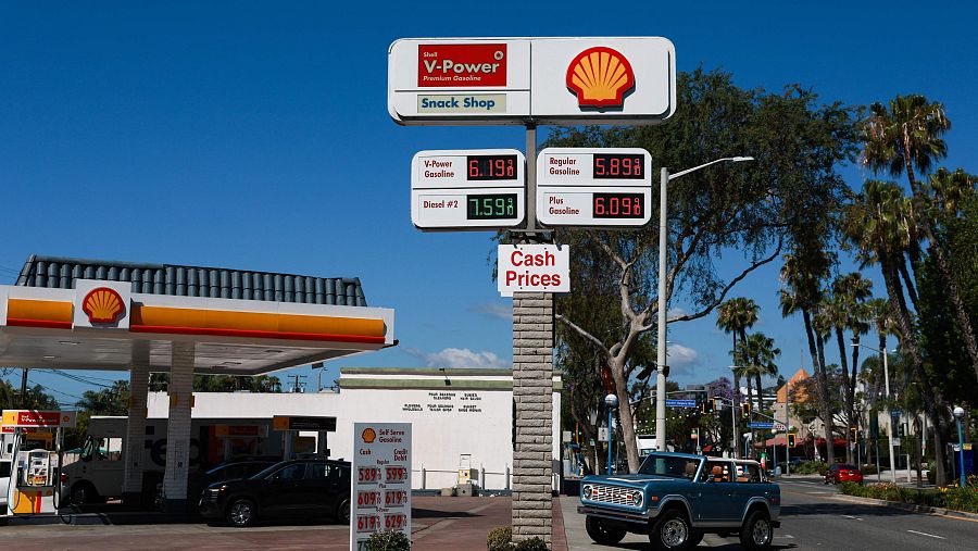 Gasolinera Shell en California con precios de gasolina y diésel visibles. Un Ford Bronco azul claro destaca en la escena junto a palmeras.