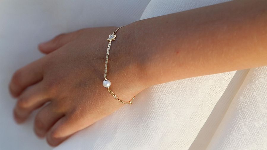 Mano de niña con pulsera dorada, perla y charm de flor con piedras. Fondo de tejido blanco.