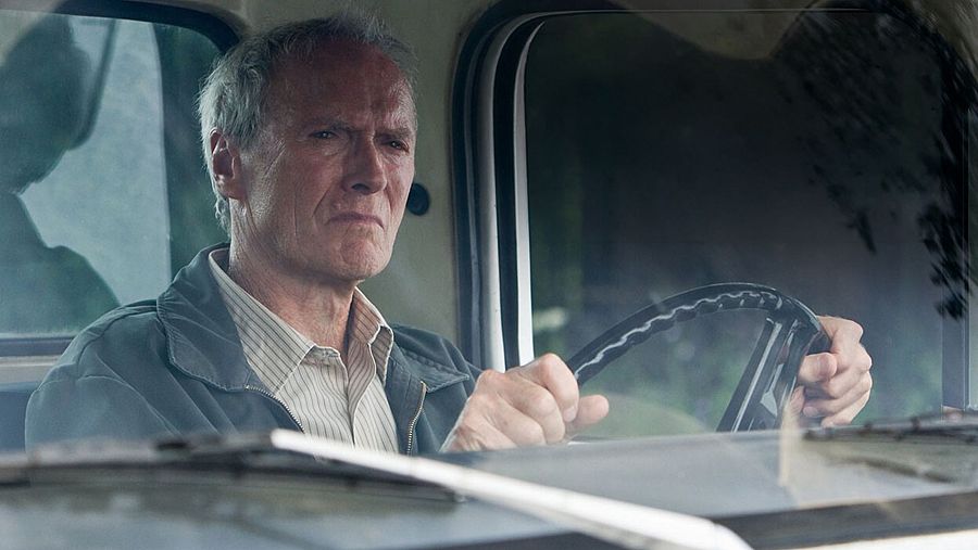 Clint Eastwood, actor de edad avanzada, conduce un vehículo con expresión seria. Viste camisa de rayas y chaqueta oscura. Se ve vegetación borrosa al exterior.