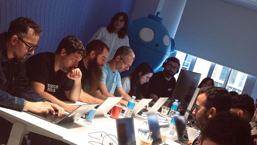 Varias personas sentdas frente a sus portátiles trabajando. De fondo, en la parte central de la fotografía se ve una especia de mascota esférica de ojos grandes.