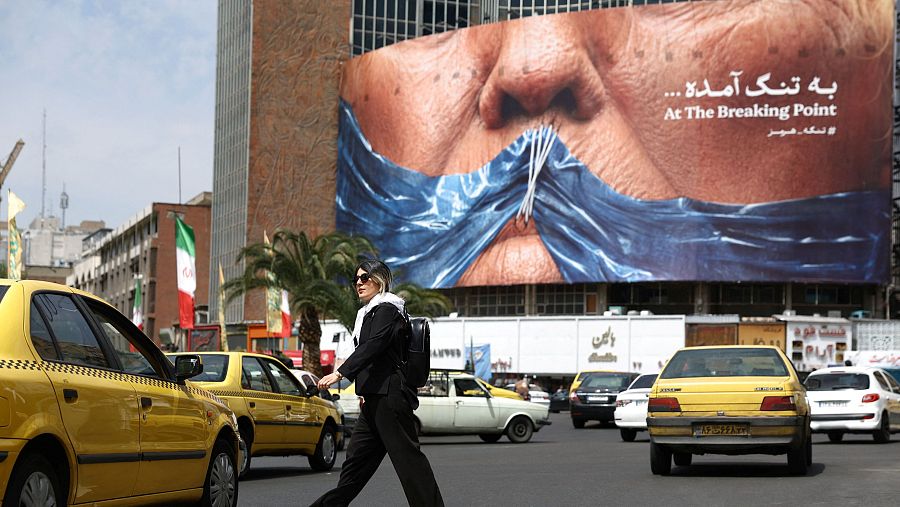 Calle de Teherán con persona en paso de cebra y cartel publicitario de rostro con boca cosida y texto 