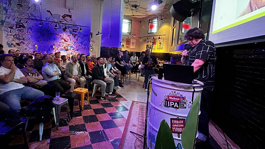 Sala de techos altos. Llena de gente sentada viendo una proyeccion. En primer término, una persona manipula un portátil colocado sobre un barril metálico