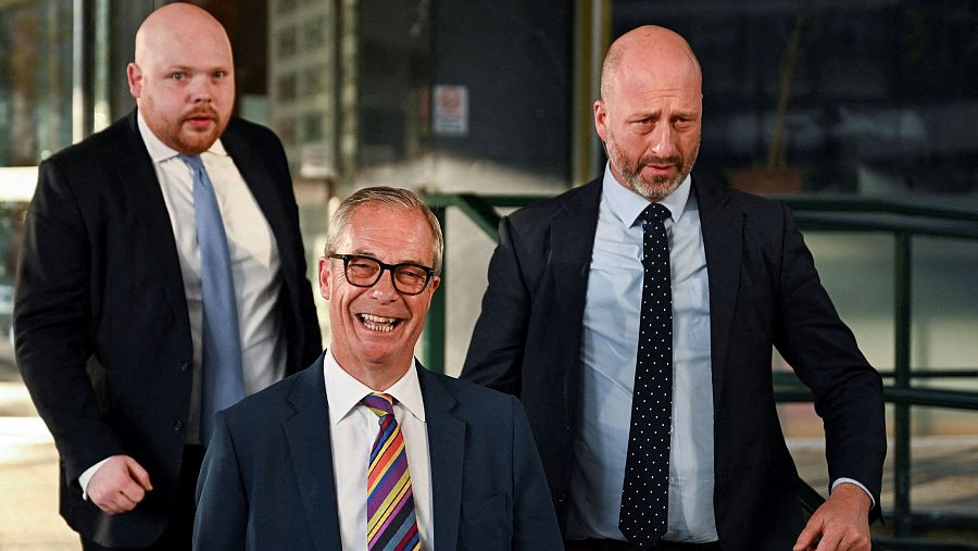 Nigel Farage, líder de Reform UK, sonríe ampliamente con traje azul y corbata colorida, flanqueado por dos hombres serios con traje y barba. Fondo arquitectónico.