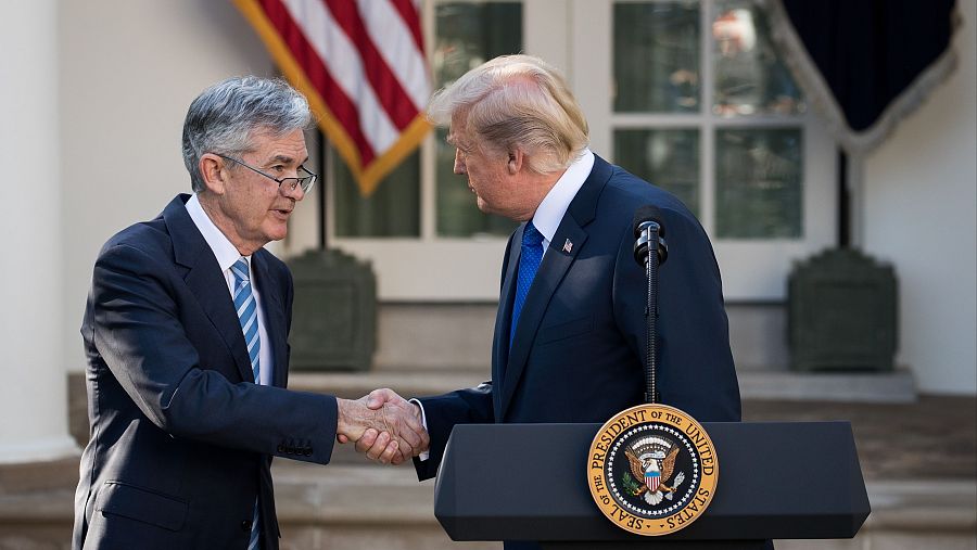 Donald Trump y Jerome Powell se dan la mano frente a un edificio blanco y una bandera de Estados Unidos. Trump está detrás de un atril con el sello presidencial.