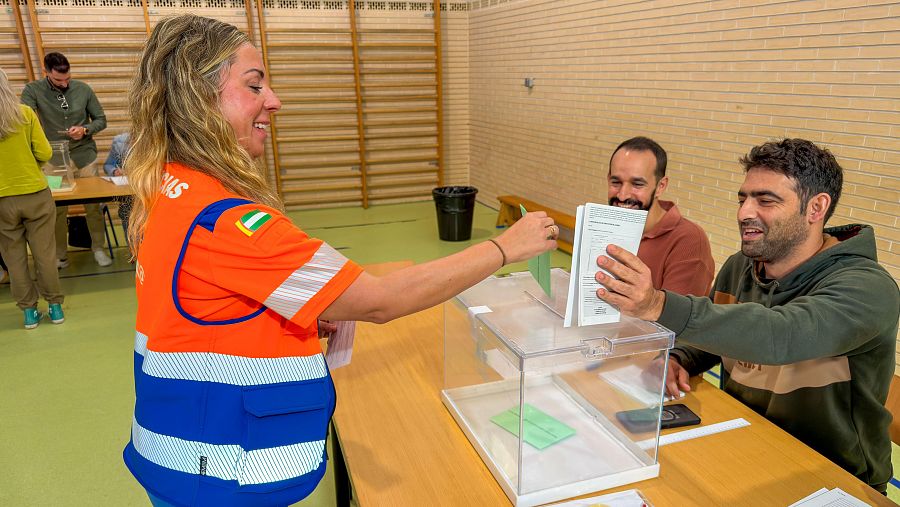 Mujer con chaleco de Andalucía introduce sobre verde en urna transparente; hombre con barba y sudadera verde oscura sostiene papeletas blancas. Entorno de colegio electoral.