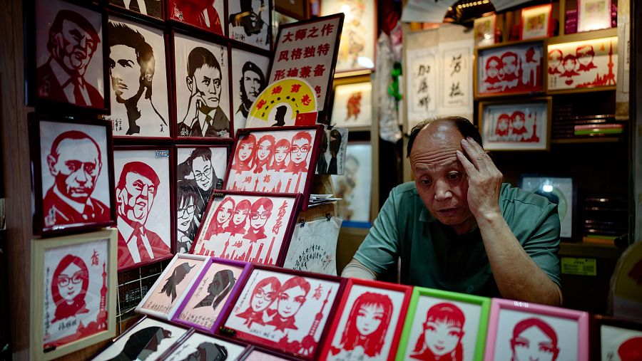 Un puesto en China exhibe retratos enmarcados de Trump, Putin, Ronaldo y Kim Jong-un, junto a un hombre mayor pensativo en primer plano.