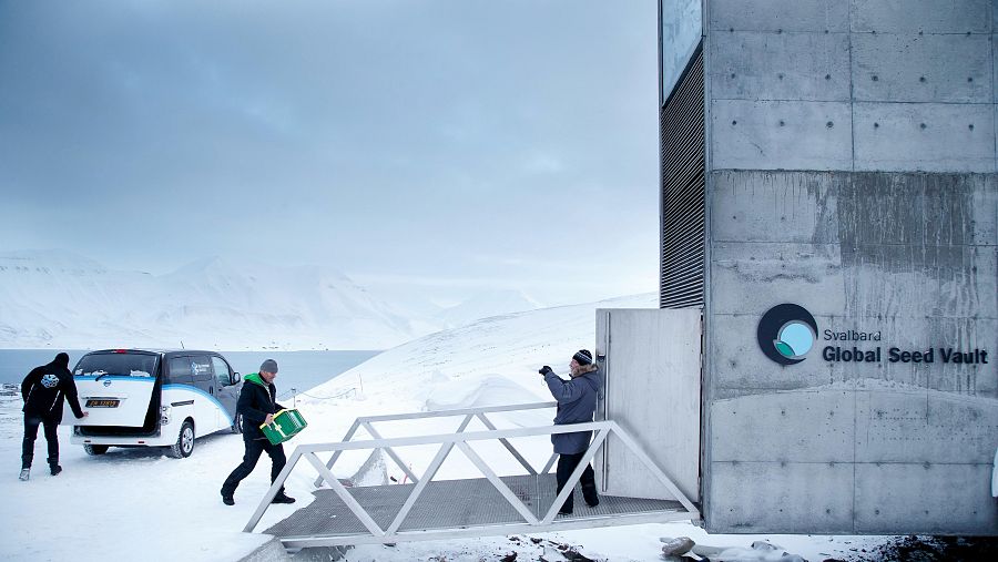 Paisaje invernal con la Bóveda Global de Semillas de Svalbard. Personas y furgoneta blanca en la nieve, una lleva un contenedor verde.