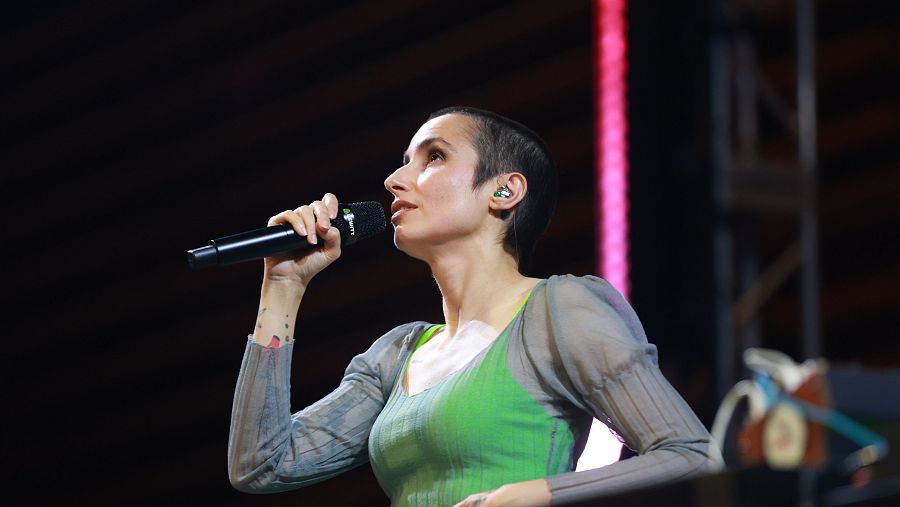 Primer plano de Zahara actuando, con pelo rapado, auricular verde y blanco, micrófono negro y tatuajes en el antebrazo. Viste camiseta verde y prenda gris semitransparente. Fondo oscuro con luces de escenario.
