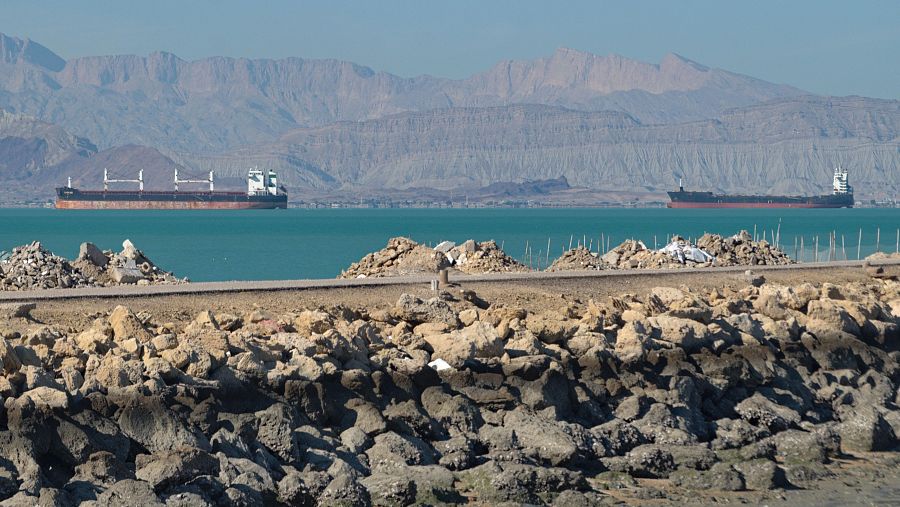 Paisaje marítimo con carguero y petrolero en aguas turquesas, montañas áridas al fondo y un terraplén de rocas en primer plano.
