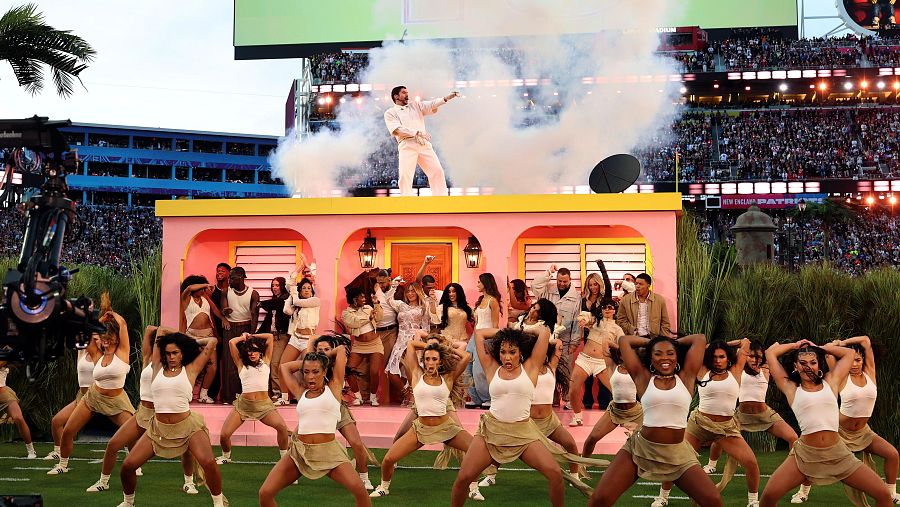 Bad Bunny canta sobre una casa rosa en un estadio, rodeado de humo y bailarines. La multitud llena las gradas del recinto.