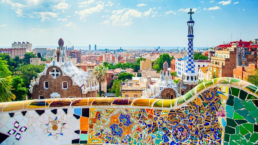 Vista panorámica del Parc Güell, Barcelona, con su banco de mosaicos, porterías de Gaudí y la ciudad al fondo bajo un cielo azul.