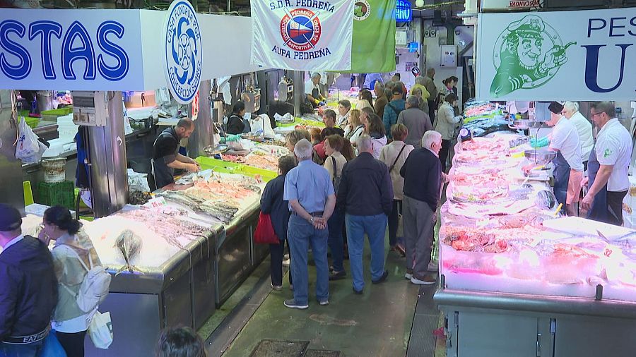 Vista aérea de un concurrido mercado de pescado interior, con puestos llenos de marisco y clientes interactuando con vendedores. Se observan carteles informativos y publicitarios.