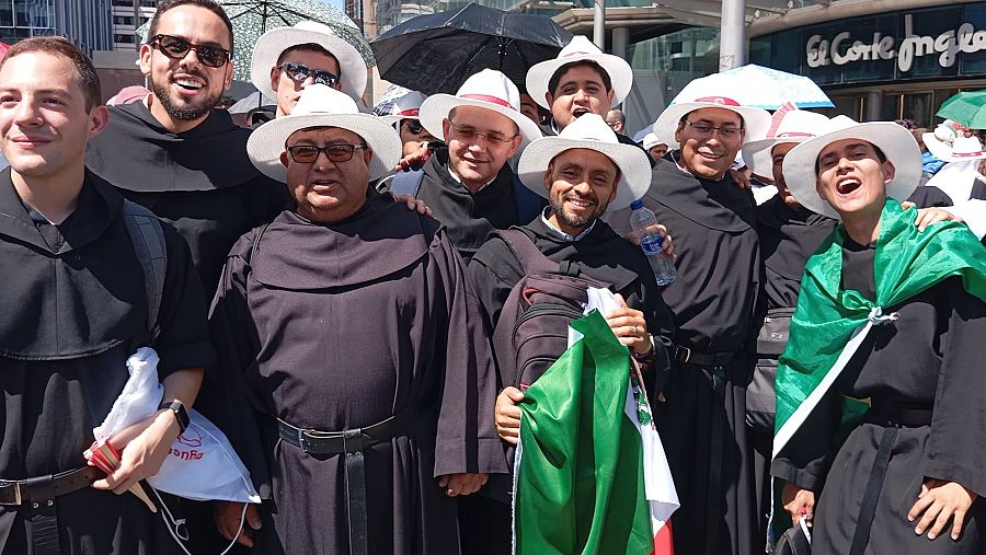 Jóvenes agustinos recoletos con hábitos y sombreros blancos, algunos portando la bandera de México, esperan en un día soleado frente a un edificio con el logo de 
