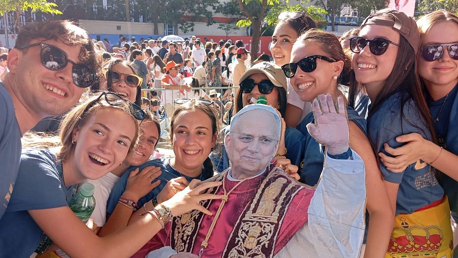 Jóvenes sonríen y posan con un recorte del Papa Juan Pablo II en un evento al aire libre en Madrid, rodeados de una multitud.