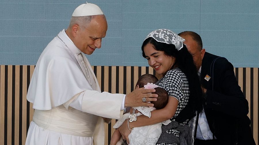 El Papa Francisco, sonriente y vestido de blanco, acaricia la cabeza de un bebé sostenido por una mujer. La escena es de gran ternura y afecto.
