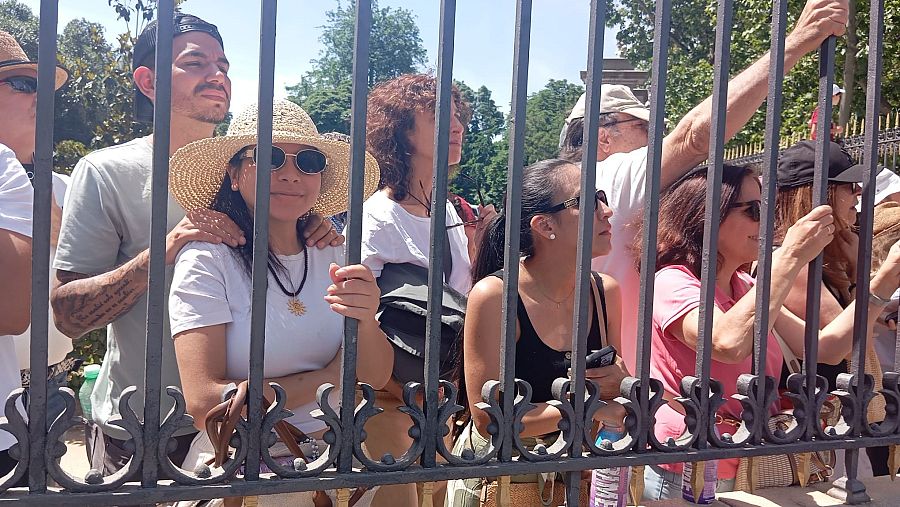 Milena, de Argentina, y su esposo observan un evento detrás de una valla de metal en un día soleado, rodeados de otras personas. Ella lleva sombrero y gafas de sol, él una gorra.
