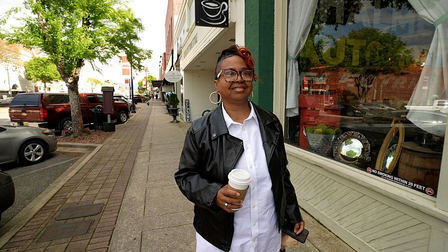 Mujer afroamericana con cabello rojizo, gafas y chaqueta de cuero, pasea por una calle con edificios, un escaparate y la señal 