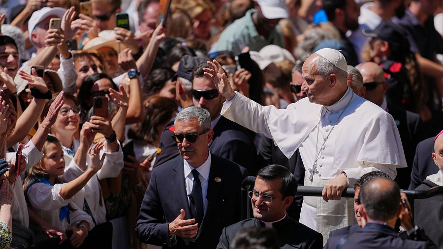El Papa Francisco saluda a una multitud eufórica, con personas levantando sus brazos y teléfonos móviles. Se observa personal de seguridad y clérigos en la escena.