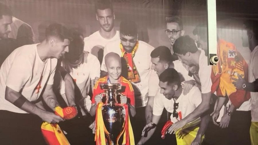 María Caamaño, sonriente y con cabeza rapada, sostiene un trofeo plateado rodeada de jugadores de fútbol, incluyendo a Sergio Busquets. Predominan blanco y negro con detalles en rojo y amarillo.