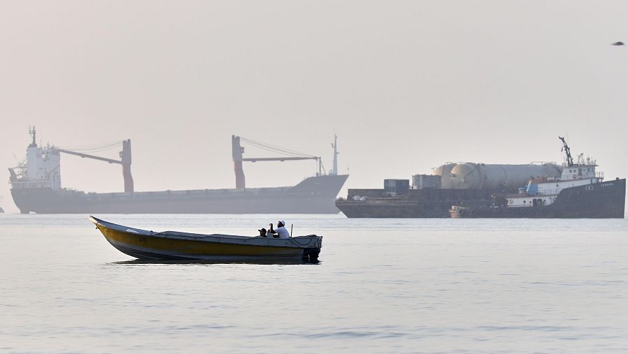 Barca amarilla y oscura con dos personas navega en aguas brumosas. Al fondo, dos buques de carga, uno con grúas rojas y otro con carga cilíndrica, se ven entre la neblina.