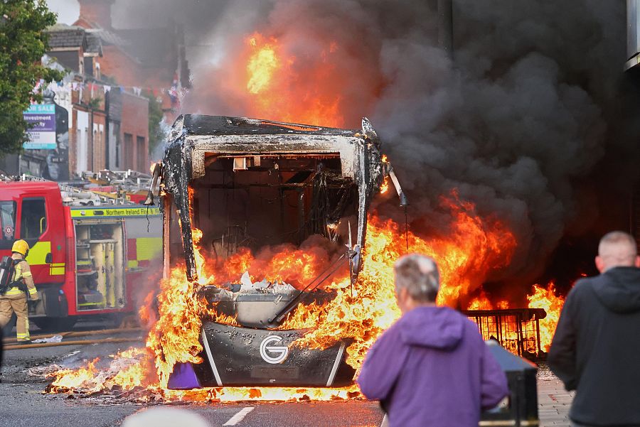 Varias personas observan cómo llegan los bomberos para apagar un vehículo que fue incendiado durante una protesta en el este de Belfast