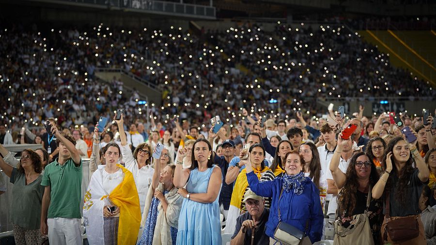 Multitud masiva en el Estadio Olímpico Lluís Companys durante una vigilia de oración nocturna, iluminada por miles de linternas de teléfonos móviles. Se observa a una mujer con túnica blanca y estola amarilla en primer plano.