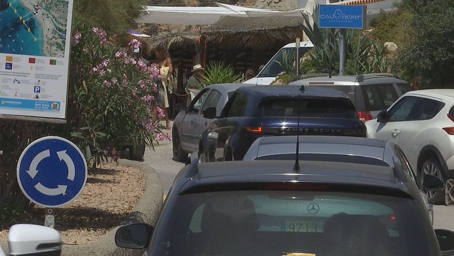 Acceso a Cala Hort Restaurant en Ibiza con señalización informativa, cola de vehículos (incluyendo Range Rover) y estructura de playa bajo un día soleado.