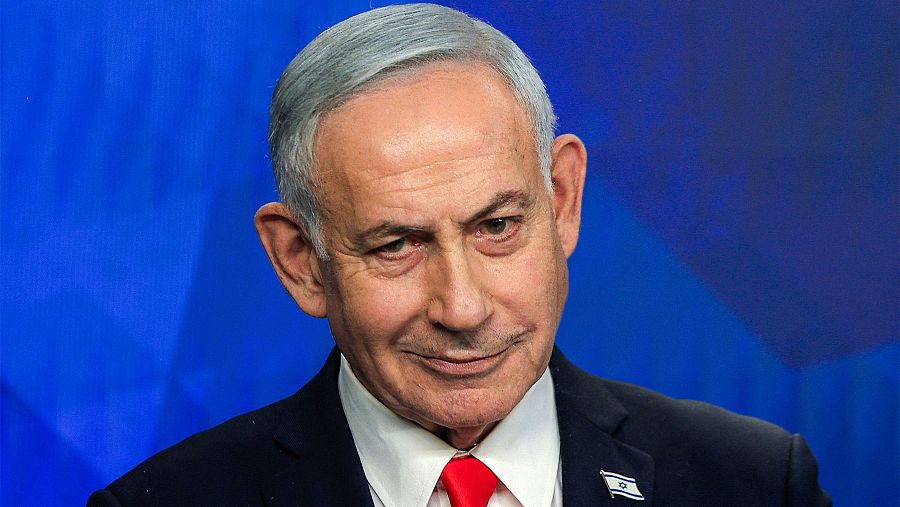 Benjamin Netanyahu, Primer Ministro de Israel, con traje oscuro, camisa blanca y corbata roja, luce un pin de la bandera israelí y una expresión pensativa.