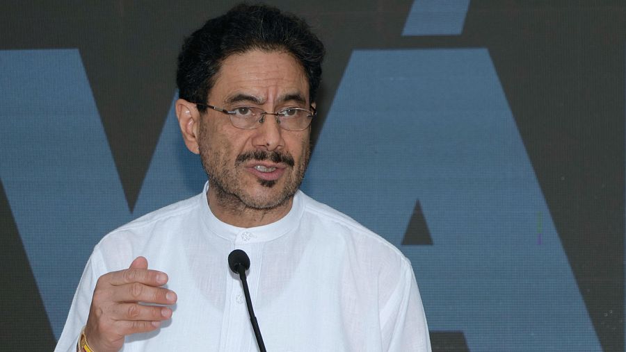 Iván Cepeda, candidato a la Presidencia de Colombia, en un primer plano con gesto de oratoria, camisa blanca y gafas. Fondo oscuro con un gran elemento gráfico azulado.