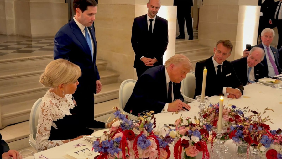 Donald Trump firma un documento en una cena diplomática nocturna, flanqueado por Emmanuel Macron y Brigitte Macron, en un elegante salón con arreglos florales.