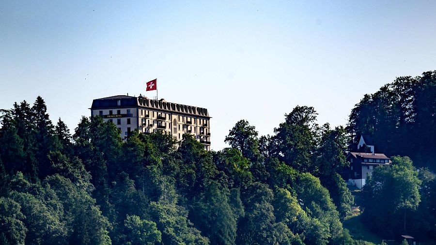 Palace Hotel sobre colina boscosa con bandera suiza. Edificio clásico de fachada clara y tejado oscuro. Cielo azul despejado.