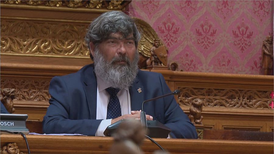 Miquel Ensenyat Riutort, político balear, con traje azul oscuro y corbata, sentado frente a un atril tallado con micrófono y una placa parcialmente visible 