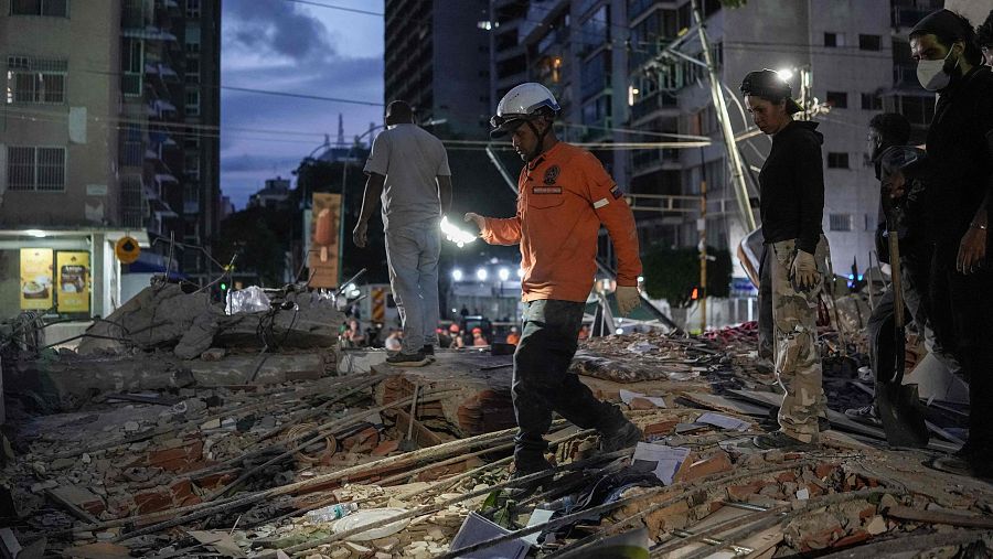 Rescatistas buscan entre escombros de un edificio derrumbado en Venezuela, iluminando la noche con linternas. Se observa un rescatista con uniforme naranja y casco blanco, y la bandera venezolana en su manga.