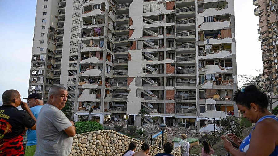 Edificio residencial blanco o gris claro gravemente dañado por un terremoto, con escombros en la base. Personas observan la escena, incluyendo un hombre con camiseta negra y roja que dice 